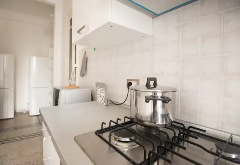 Bright, modern kitchen corner with a gas hob and a stainless-steel pot on the burner; clean countertop, tiled backsplash and appliances visible in the background.