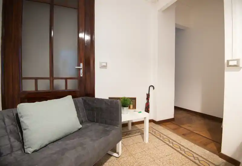 Compact, cozy living area with a dark gray upholstered sofa, pale green cushion and a small white side table with a plant; warm wooden door and patterned floor tiles add character.