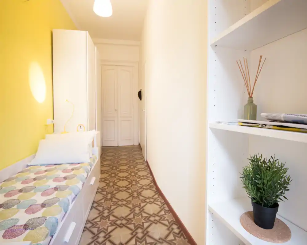 Small bright bedroom with a single bed along a yellow wall, patterned tiled floor and open shelving with decorative plants — compact and welcoming single-room layout ideal for short stays.