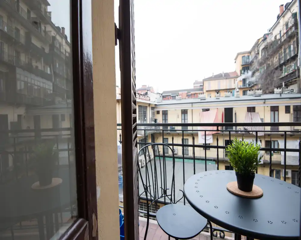 Small private balcony with a round table, two metal chairs and a potted plant, overlooking neighboring courtyard buildings — ideal for morning coffee.