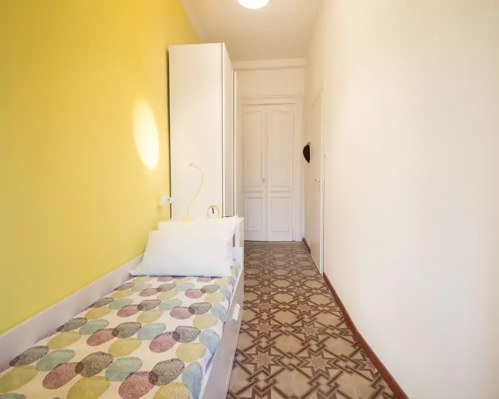 Compact single bedroom with a patterned single bed alongside a bright yellow accent wall, neat bedside lamp and wardrobe at the end of the room. The space is bright and clean, suitable for a cozy solo stay.