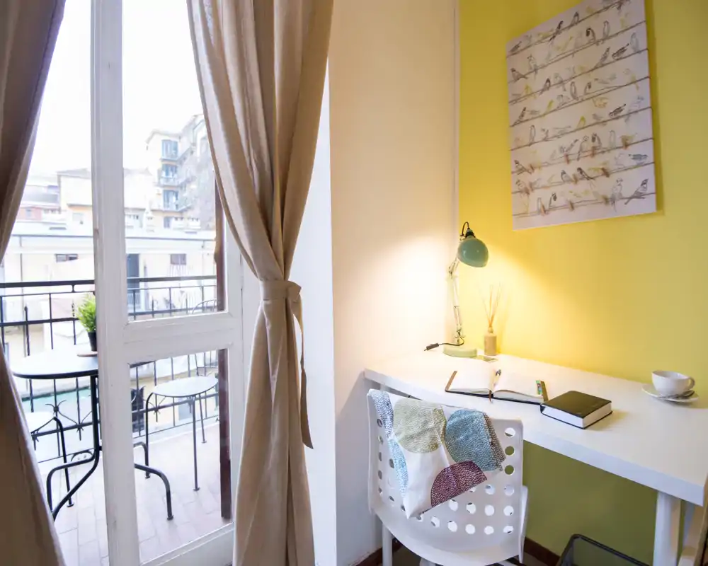 Bright corner workspace with a white desk, chair and a green desk lamp, opening onto a small balcony; warm yellow wall and decorative print create a cozy feel.