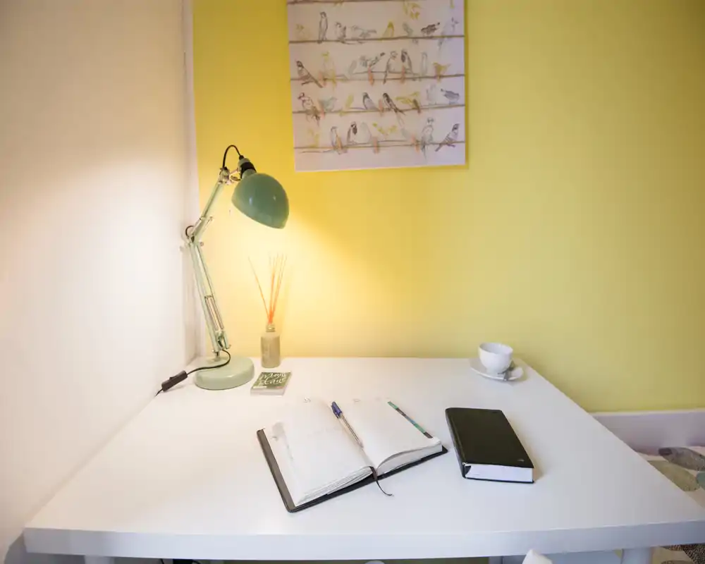 Bright, minimal bedside/desk area in a bedroom with a lamp, open notebook and cup — a tidy workspace adjacent to the bed visible at the frame edge.