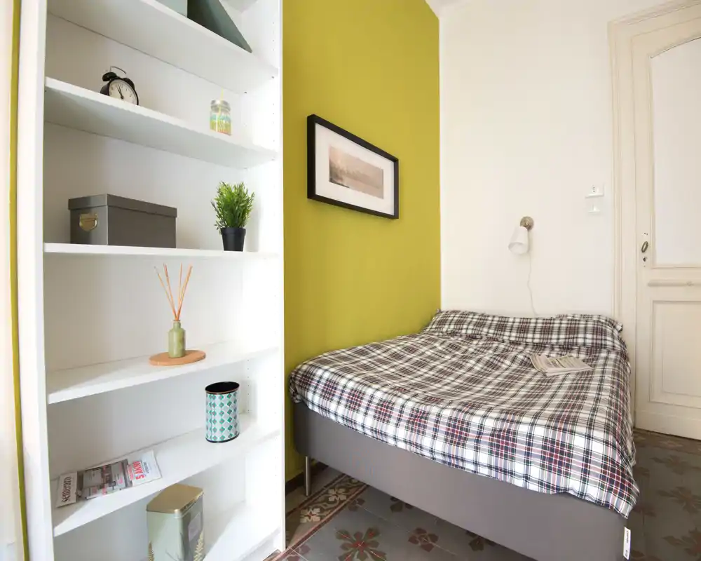 Camera da letto piccola e luminosa con letto matrimoniale coperto da una trapunta a quadri, parete verde a contrasto e scaffalatura ordinata—accogliente e fotografata per affitti.