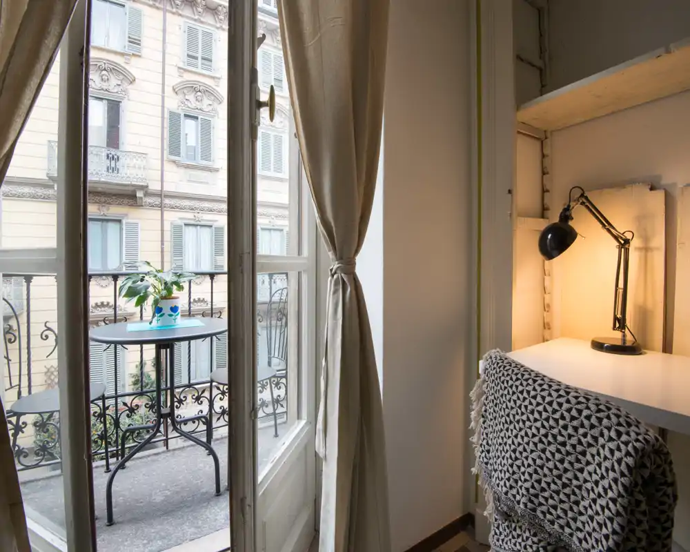 Zona giorno luminosa con un piccolo tavolo sul balcone visibile attraverso le porte-finestre, tende morbide e un angolo scrivania accogliente con lampada — ideale per colazione o lavoro alla luce naturale.