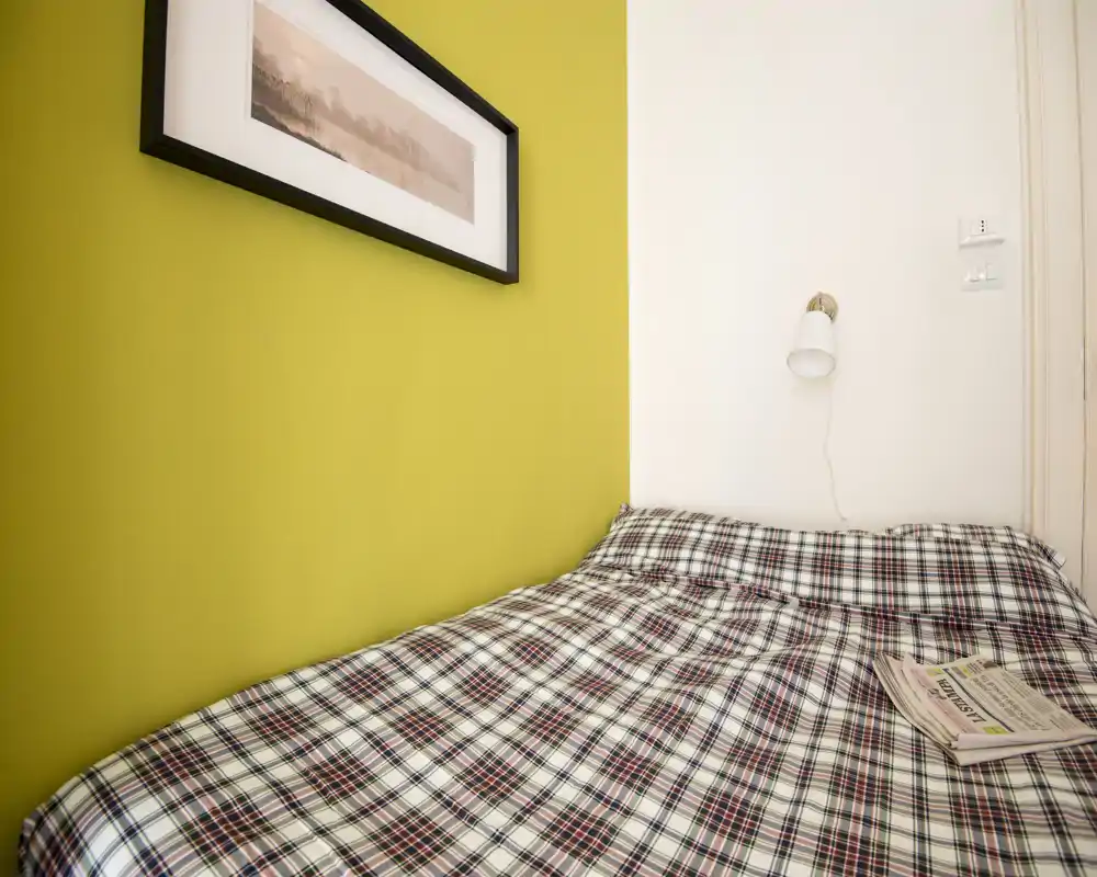 Camera da letto compatta e luminosa con piumone a quadri, lampada da parete e una parete d'accento verde — ideale per una persona o soggiorni brevi.