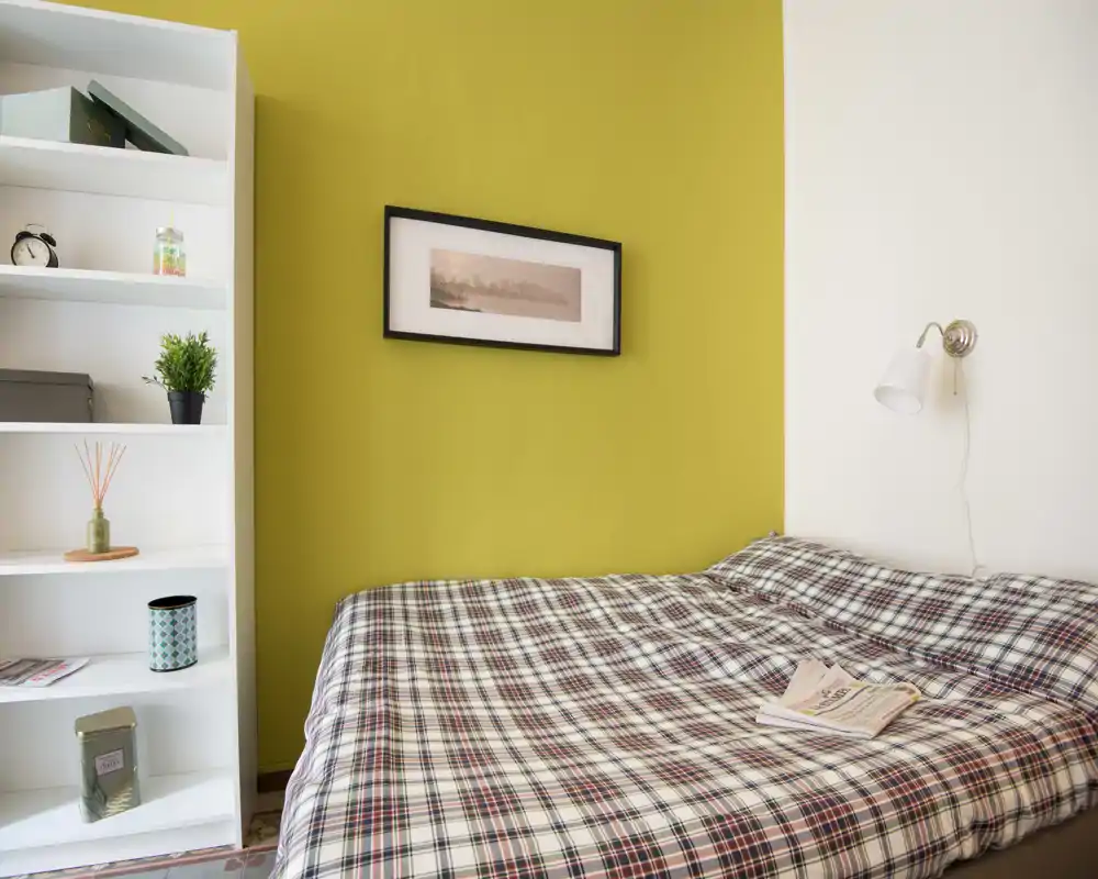 Camera da letto luminosa e accogliente con copriletto a quadri, lampada da lettura a muro e quadro su una parete verde; scaffale ordinato per riporre oggetti.