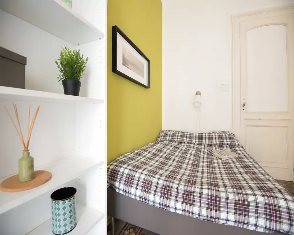 Camera da letto compatta e luminosa con un letto matrimoniale a fantasia appoggiato contro una parete verde chiaro, lampada da lettura a parete e mensole decorative in primo piano.