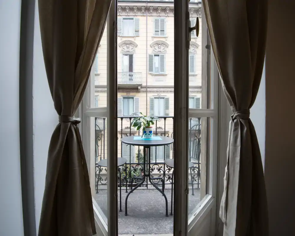 Piccolo balcone cittadino visto attraverso porte finestre aperte, con un tavolino da bistrò, due sedie e una pianta in vaso; si intravede la facciata classica dell'edificio di fronte.