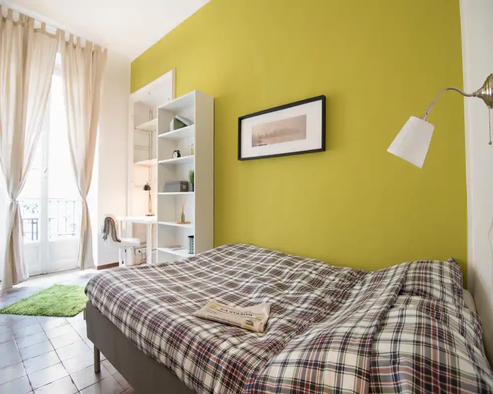 Camera da letto luminosa e accogliente con un ampio letto matrimoniale con copriletto a quadri, parete accentuata verde, libreria e angolo scrivania vicino a una finestra alta che illumina la stanza.