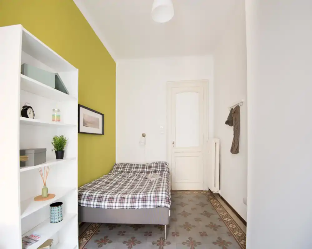 Camera da letto luminosa e semplice con letto matrimoniale contro una parete verde chiaro, piastrelle a motivo e una libreria aperta per riporre oggetti e decorazioni.