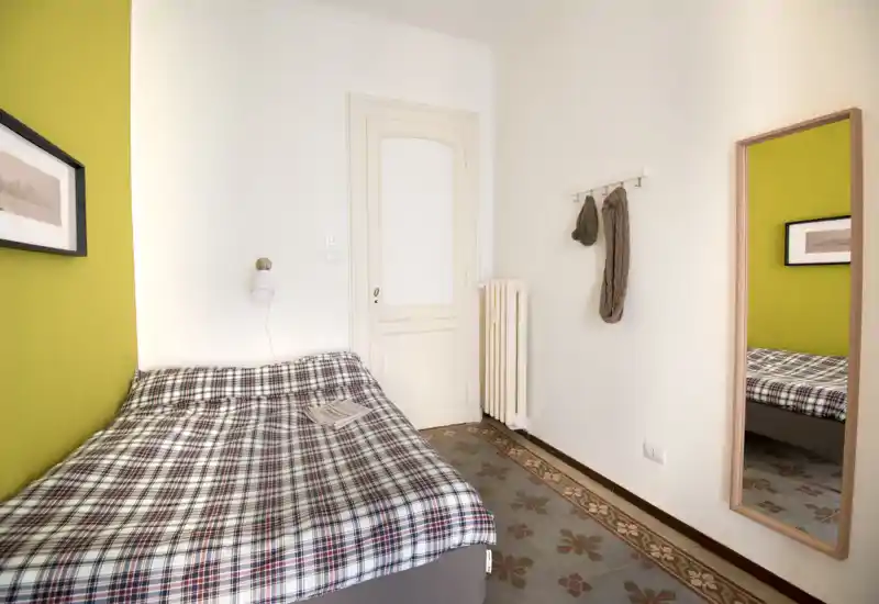 Camera singola accogliente con copriletto a quadri, pareti luminose e pavimento piastrellato decorativo. Uno specchio a figura intera e dei ganci a parete aggiungono funzionalità, la stanza è ben illuminata.