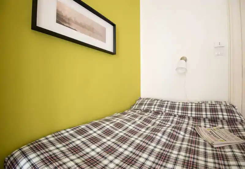 Camera da letto compatta e luminosa con piumone a quadri, lampada da parete e una parete d'accento verde — ideale per una persona o soggiorni brevi.
