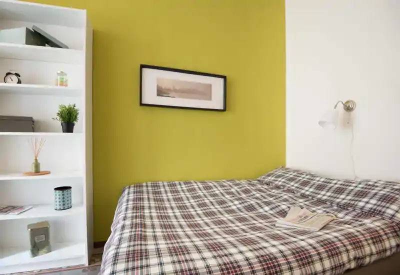 Camera da letto luminosa e accogliente con copriletto a quadri, lampada da lettura a muro e quadro su una parete verde; scaffale ordinato per riporre oggetti.