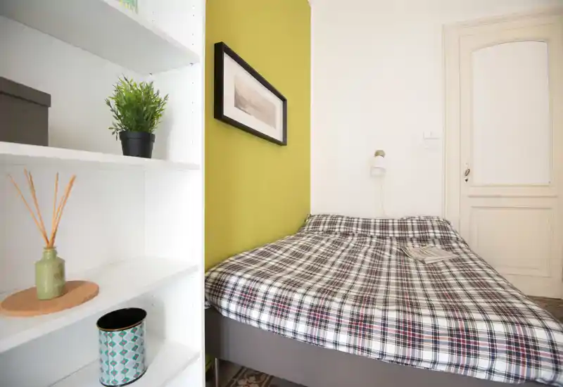 Camera da letto compatta e luminosa con un letto matrimoniale a fantasia appoggiato contro una parete verde chiaro, lampada da lettura a parete e mensole decorative in primo piano.