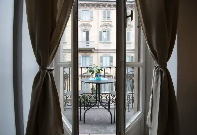 Piccolo balcone cittadino visto attraverso porte finestre aperte, con un tavolino da bistrò, due sedie e una pianta in vaso; si intravede la facciata classica dell'edificio di fronte.