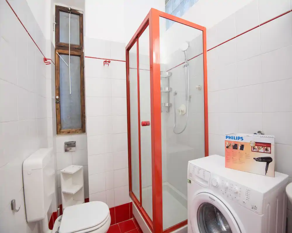 Bagno compatto con box doccia in vetro con cornice rossa, WC e lavatrice; pareti piastrellate bianche e pavimento rosso che rendono l'ambiente luminoso e pulito.
