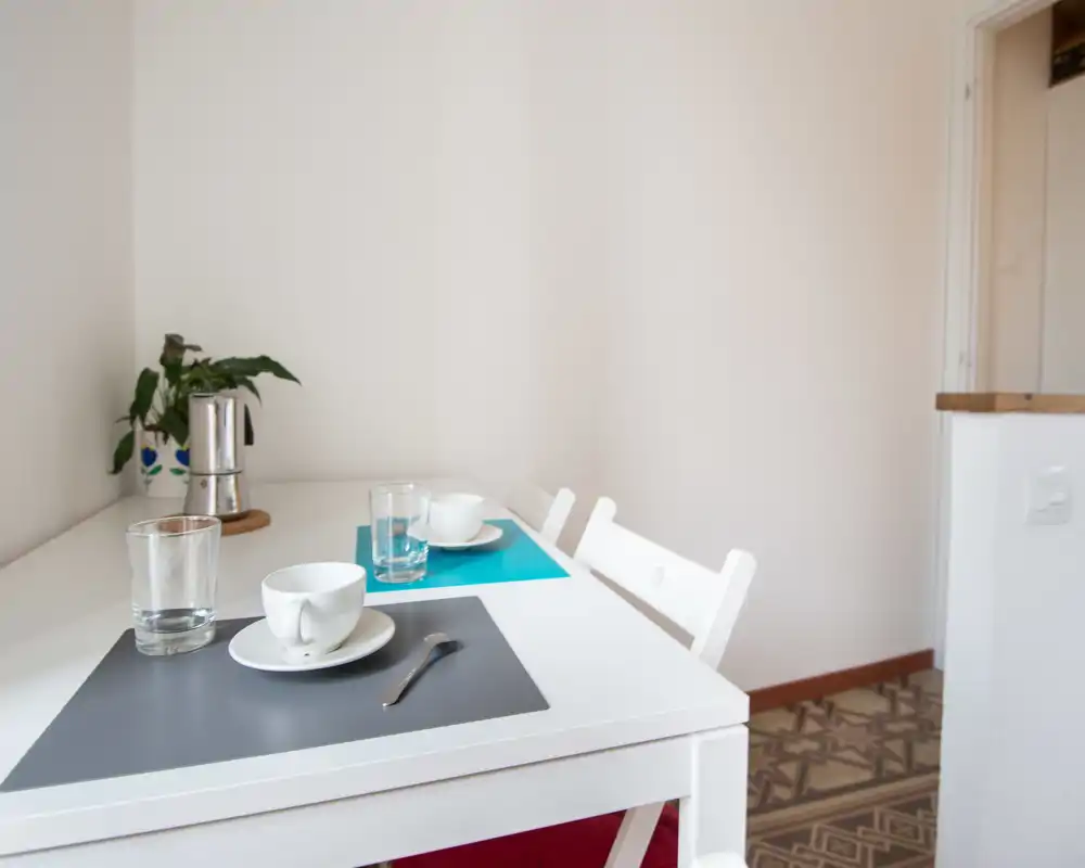 Zona pranzo minimal e luminosa con tavolo bianco apparecchiato per due, tovagliette colorate, bicchieri e una piccola pianta — un angolo colazione semplice e accogliente.