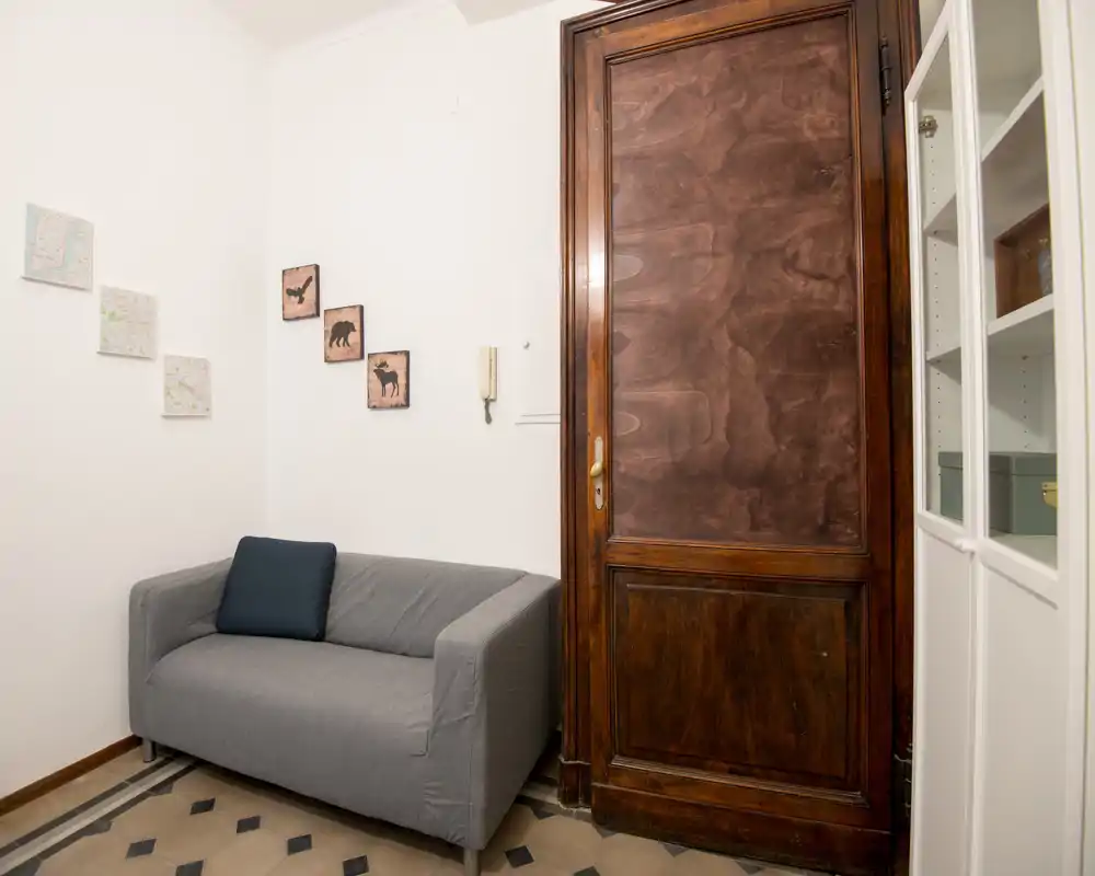 Angolo living compatto con divano grigio a due posti, quadretti decorativi e una grande porta in legno — un angolo ordinato e illuminato, adatto a leggere o rilassarsi.