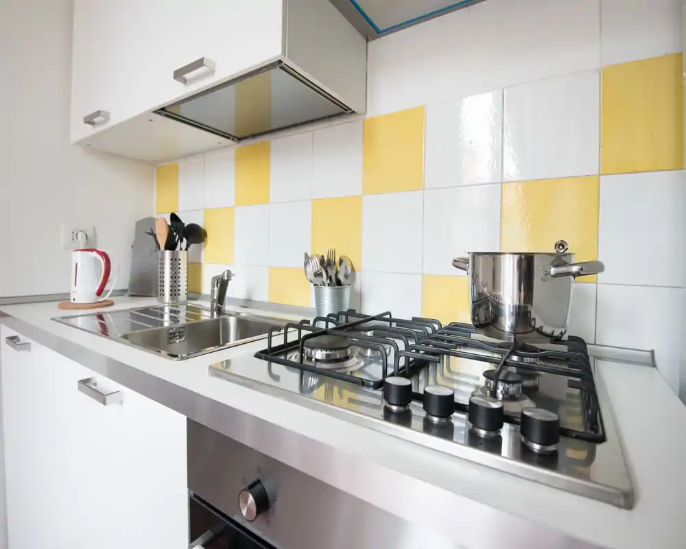 Cucina compatta e moderna con piano cottura a gas in acciaio, lavello e paraschizzi a piastrelle gialle e bianche; luminosa e ordinata, ideale per affitti brevi.