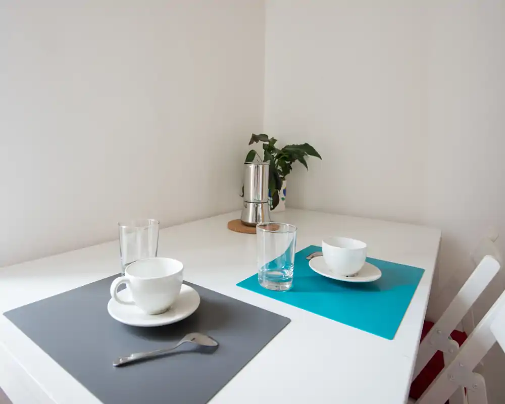 Angolo colazione luminoso e minimale con due coperti su un tavolo bianco, tazze semplici, bicchieri e una piccola pianta — ideale per un angolo pranzo compatto.