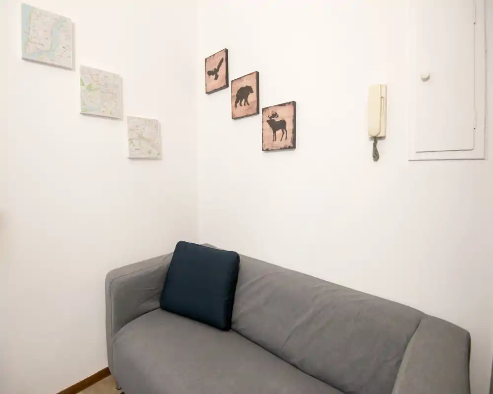 Zona giorno compatta con divano grigio per due persone, cuscino scuro e quadri minimal alle pareti: ambiente semplice, pulito e ben illuminato.