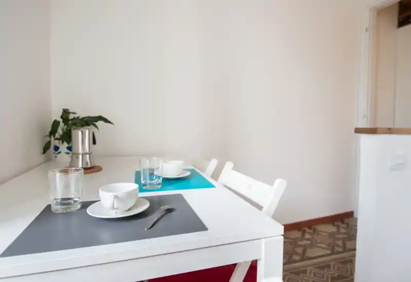 Zona pranzo minimal e luminosa con tavolo bianco apparecchiato per due, tovagliette colorate, bicchieri e una piccola pianta — un angolo colazione semplice e accogliente.