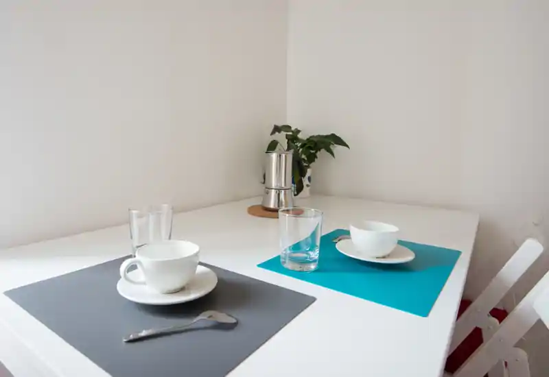 Angolo colazione luminoso e minimale con due coperti su un tavolo bianco, tazze semplici, bicchieri e una piccola pianta — ideale per un angolo pranzo compatto.
