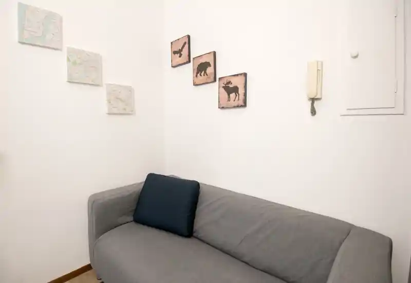Zona giorno compatta con divano grigio per due persone, cuscino scuro e quadri minimal alle pareti: ambiente semplice, pulito e ben illuminato.