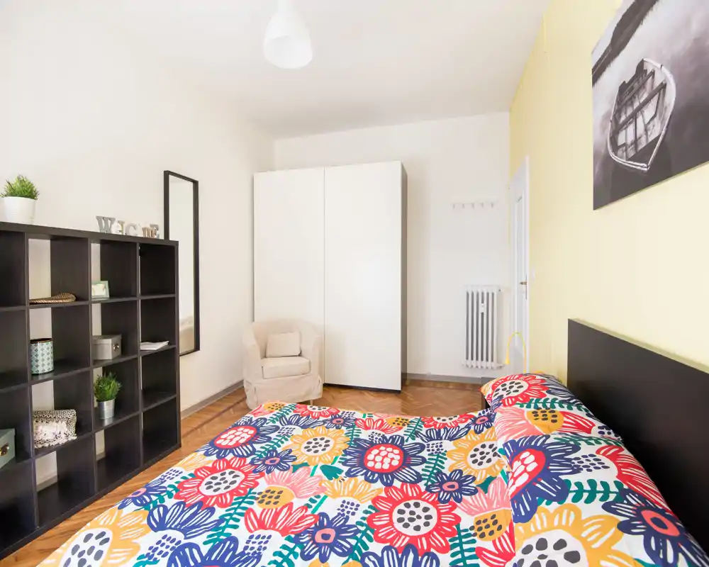 Dormitorio luminoso y ordenado con edredón floral colorido, armario, estantería y un sillón acogedor — estilo limpio y buena luz natural.