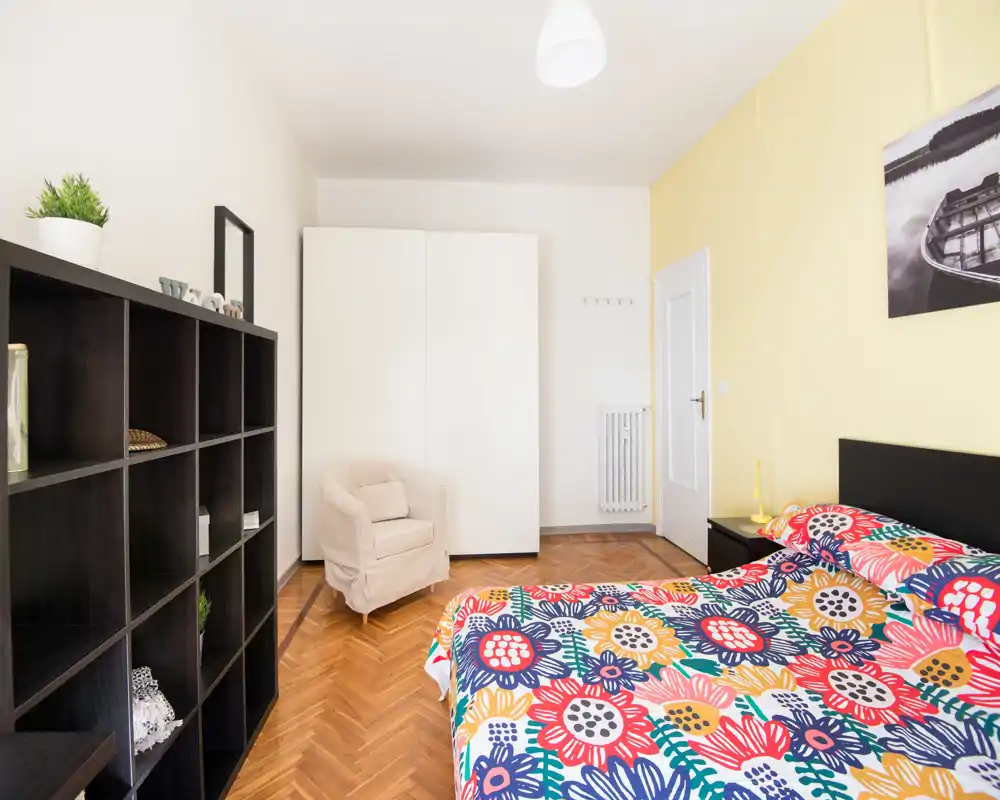 Dormitorio luminoso y bien decorado con edredón floral colorido, mesita de noche, armario y un sillón cómodo. Perfecto para anuncios de alquiler que destaquen la decoración alegre y el espacio de almacenamiento.