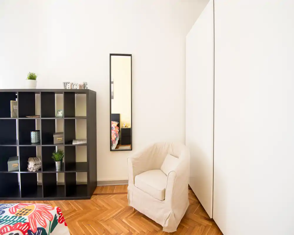 Rincón de dormitorio luminoso y minimalista con un cómodo sillón, estantería de cubos y un vislumbre de una colcha colorida reflejada en un espejo alto. El espacio es aireado y bien iluminado, ideal para estancias acogedoras.