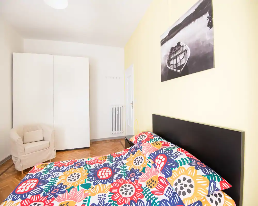 Dormitorio luminoso y ordenado con colcha floral de colores, cabecero negro y armario blanco. La habitación está bien iluminada y resulta acogedora, con un sillón en la esquina.