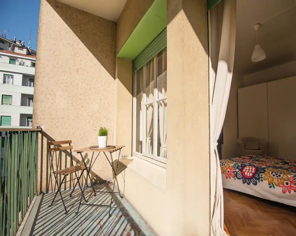 Dormitorio luminoso con acceso a un balcón privado. Se ve una cama colorida y una pequeña mesa exterior bañadas por luz natural.