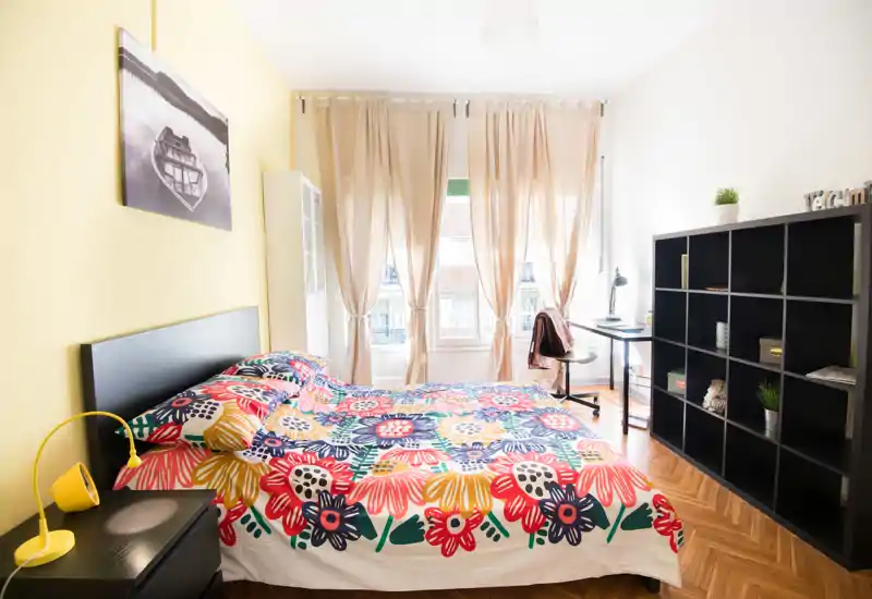 Dormitorio luminoso y bien decorado con una cama doble grande cubierta con una colcha floral colorida, mesita con lámpara, estantería y un escritorio junto a la ventana que aporta abundante luz natural.