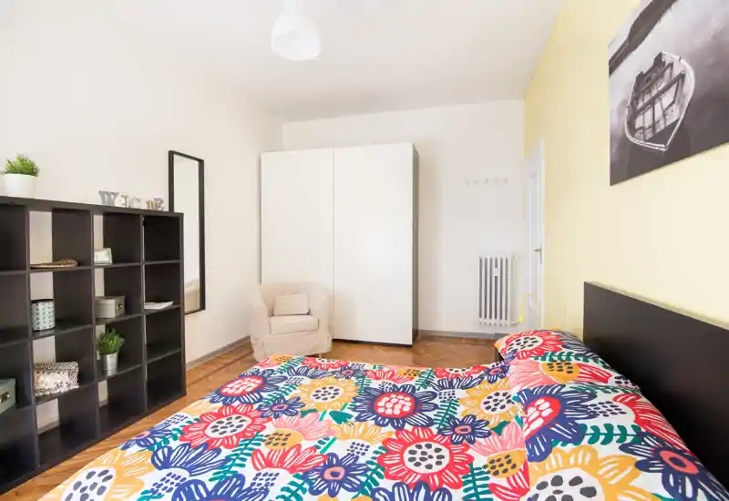 Dormitorio luminoso y ordenado con edredón floral colorido, armario, estantería y un sillón acogedor — estilo limpio y buena luz natural.
