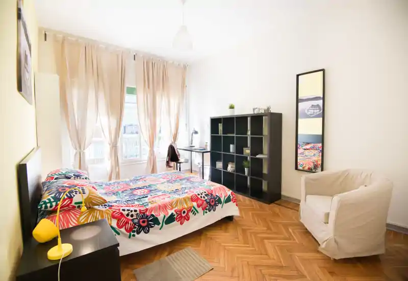 Dormitorio luminoso y bien decorado con cama doble con ropa de cama floral, mesita con lámpara, un sillón acogedor y un escritorio junto a grandes ventanas con abundante luz natural.