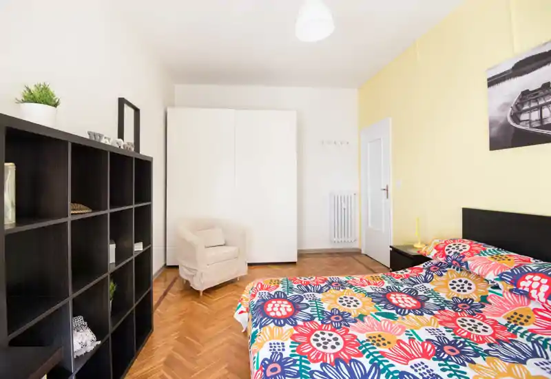 Dormitorio luminoso y bien decorado con edredón floral colorido, mesita de noche, armario y un sillón cómodo. Perfecto para anuncios de alquiler que destaquen la decoración alegre y el espacio de almacenamiento.