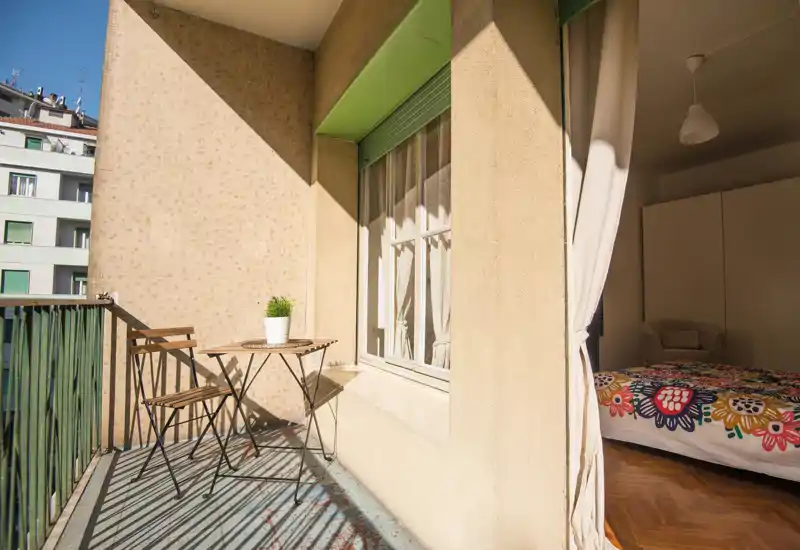 Camera da letto luminosa con accesso a un balcone privato. Si vede un letto colorato e un piccolo tavolino all'esterno illuminati da luce naturale calda.