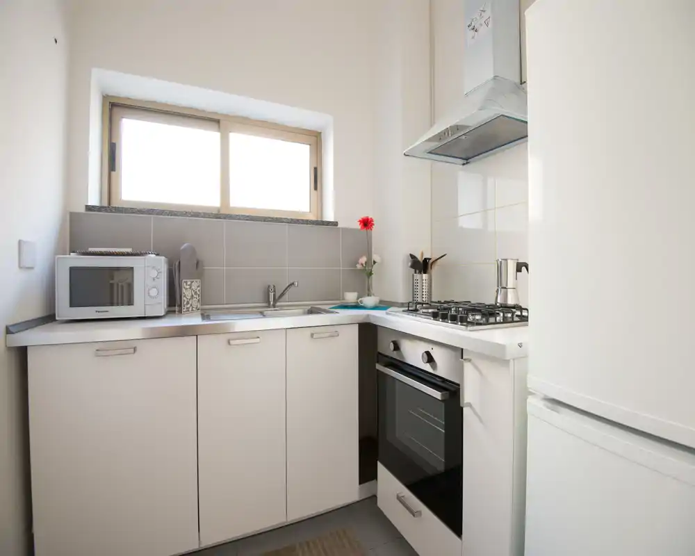 Cocina compacta y luminosa con placa de gas, horno empotrado, fregadero y pequeños electrodomésticos. La luz natural desde la ventana realza los muebles blancos y la encimera ordenada.