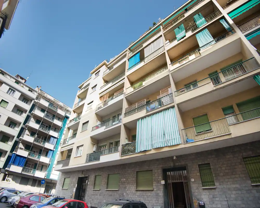 Vista exterior de un edificio residencial de varias plantas con múltiples balcones y coches aparcados en la calle, tomada en un día soleado.
