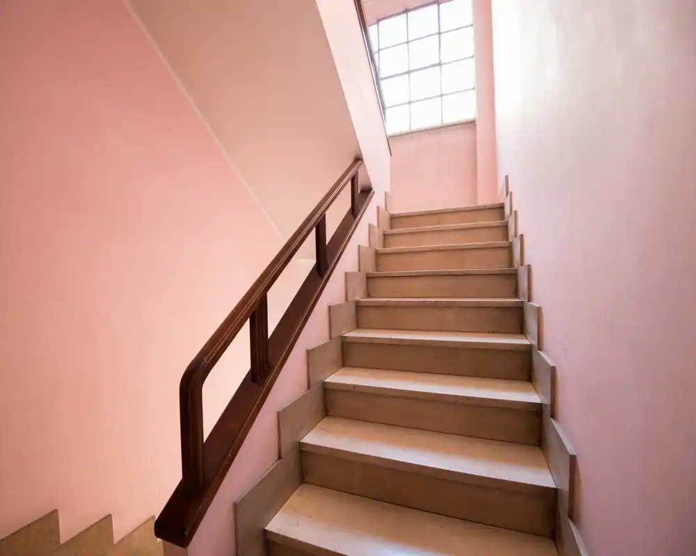 Escalera interior bien iluminada con paredes rosa pálido y pasamanos de madera que conduce a un rellano con ventana.