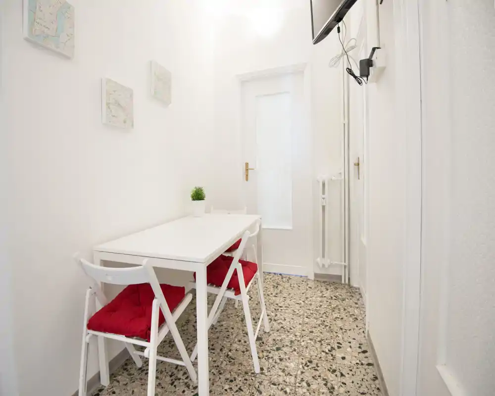 Pequeña zona de comedor con mesa blanca y sillas plegables con cojines rojos, paredes luminosas y suelo de terrazo — ideal para comidas informales.