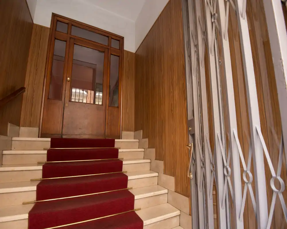 Entrada clásica de edificio con escalera de mármol alfombrada en rojo, paredes de madera y reja metálica lateral — vestíbulo luminoso y bien cuidado ideal para anuncios.