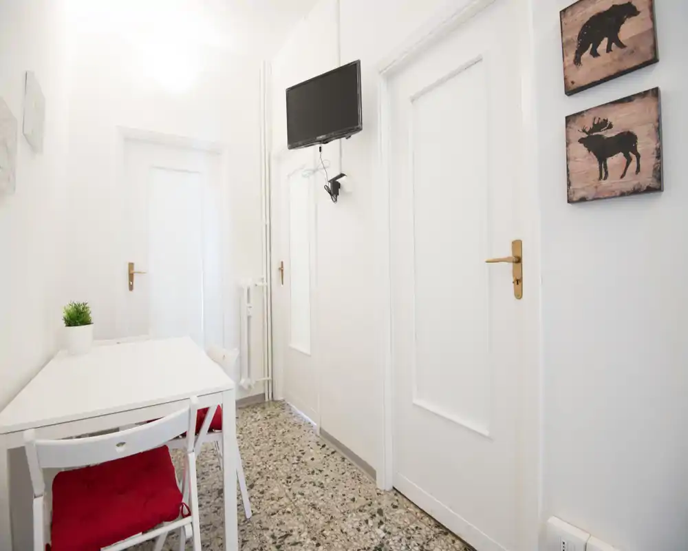 Entrada/pasillo compacto y luminoso con paredes blancas, suelo de terrazo y una pequeña mesa con sillas; televisor en la pared y placas decorativas que crean un espacio ordenado y acogedor.