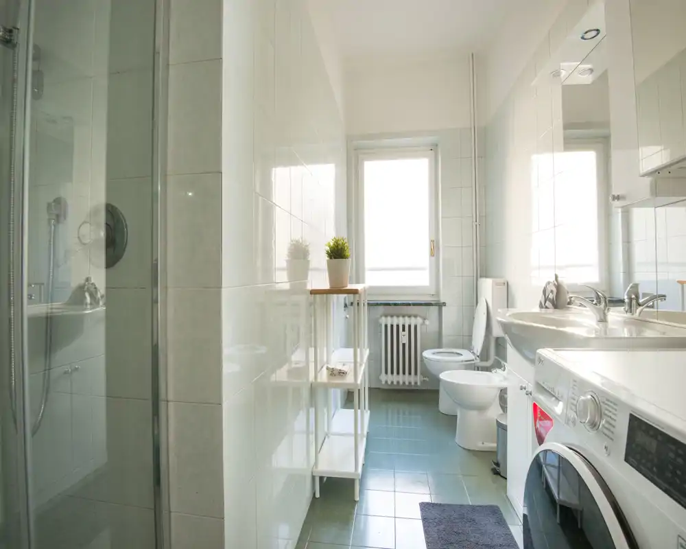 Baño luminoso y moderno con ducha de cristal, inodoro, bidé, lavabo con espejo y lavadora de carga frontal; paredes alicatadas y ventana que aporta luz natural.