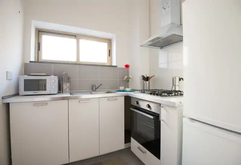 Cucina compatta e luminosa con piano cottura a gas, forno incassato, lavello e piccoli elettrodomestici. La luce naturale dalla finestra valorizza le ante bianche e il piano di lavoro ordinato.