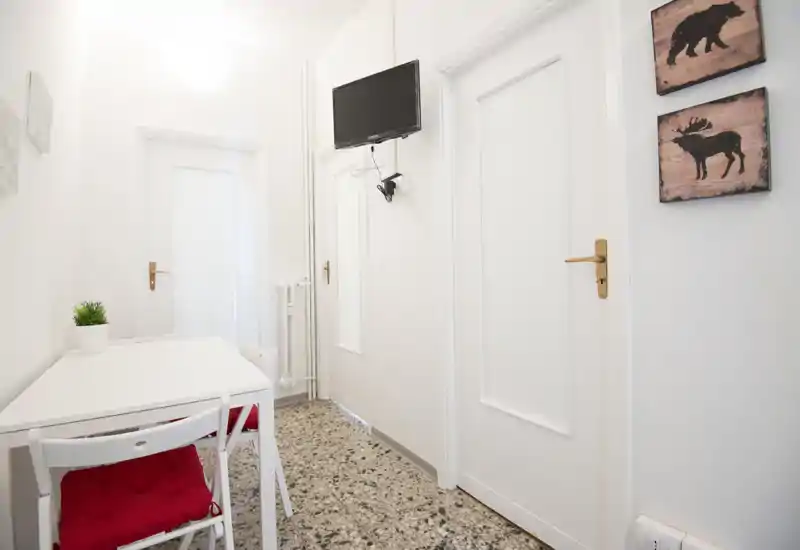 Ingresso/corridoio compatto e luminoso con pareti bianche, pavimento in graniglia e un piccolo tavolo con sedie; TV a parete e pannelli decorativi donano un aspetto ordinato e accogliente.