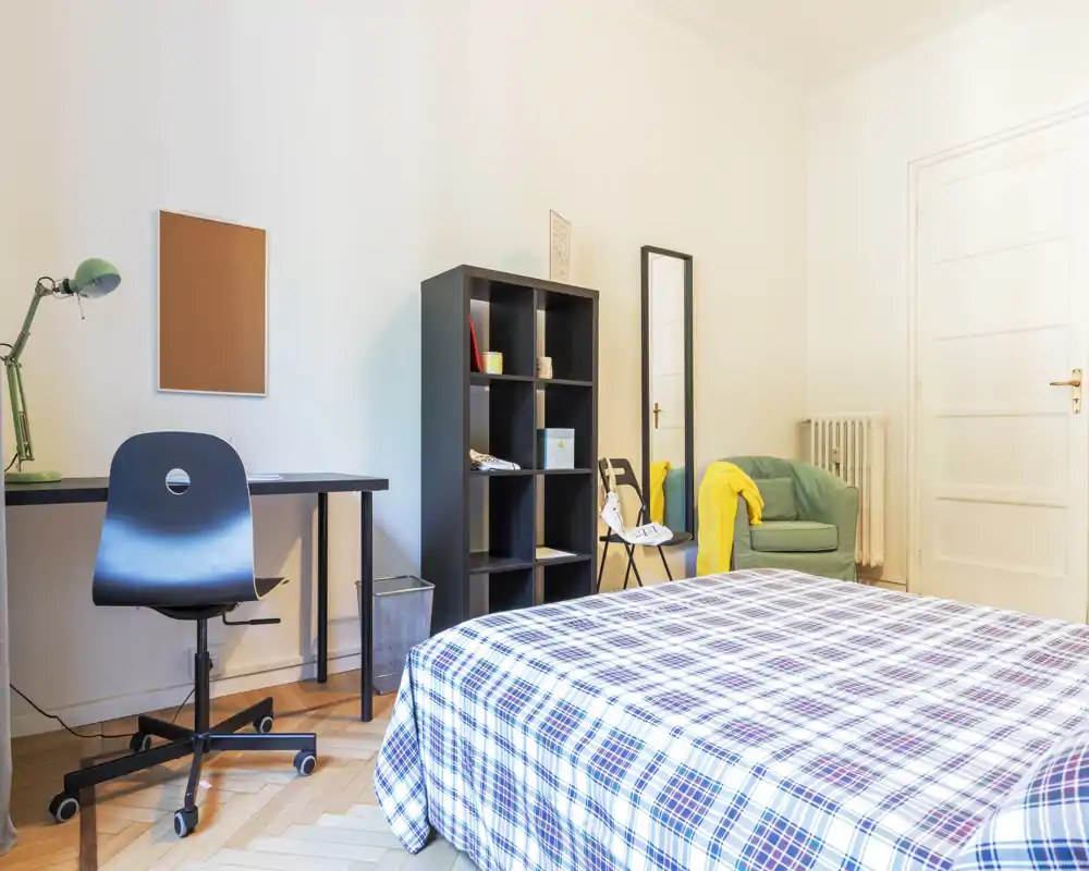Dormitorio luminoso y ordenado con una cama grande estampada en primer plano, escritorio y silla a la izquierda, estantería y butaca acogedora con una manta a la derecha — ideal para una persona o estudiante.