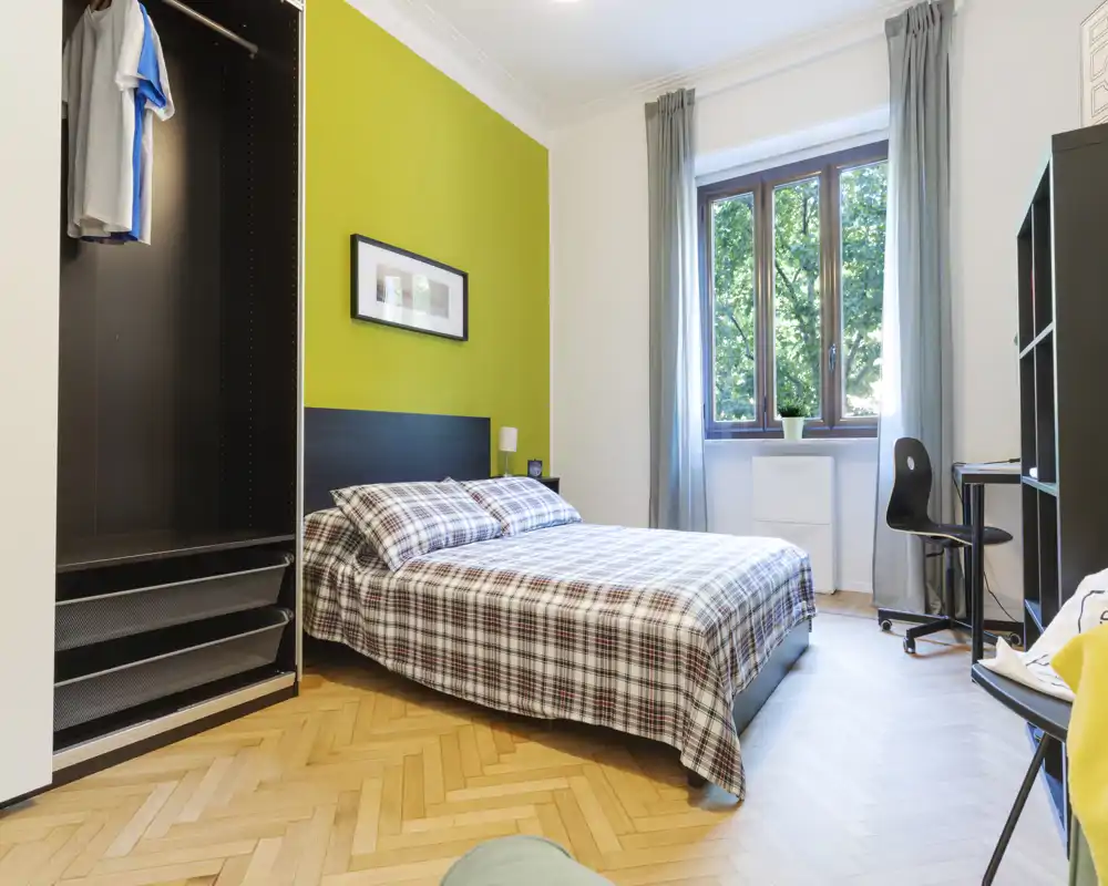 Dormitorio luminoso y ordenado con cama doble con ropa a cuadros, pared verde de acento, armario y escritorio junto a la ventana — ideal para una estancia cómoda.